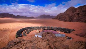 A desert landscape with a large, circular structure surrounded by tents and vehicles, suggesting a temporary accommodation setup. - Desert Sand Camp (Wadi Rum)