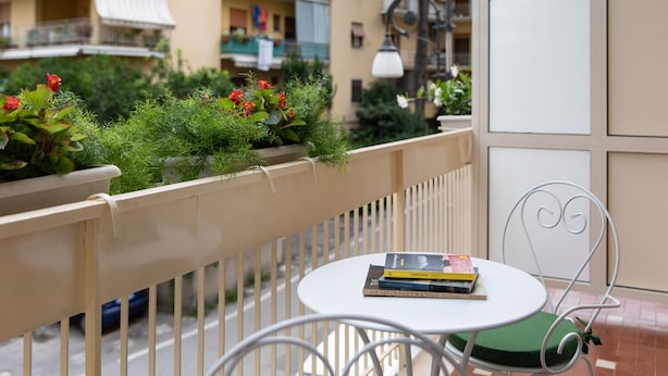 A balcony with a white metal chair and a small table, a potted plant with red flowers, and a view of a yellow building with balconies. - VIAPARSANO - two bedrooms flat (Sorrento)