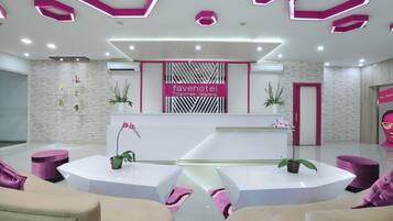 Uma área de recepção de hotel moderna, com um balcão de atendimento branco, assentos roxos e plantas decorativas.