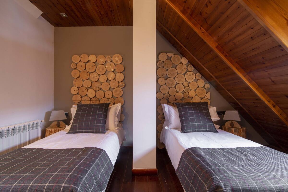 Quarto com duas camas, cada uma com cabeceira de toras empilhadas, roupa de cama branca e cobertores de retalhos xadrez.