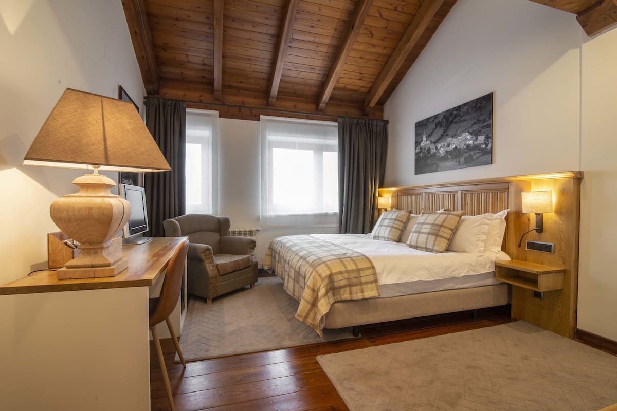 Quarto com teto de madeira, cama com colcha xadrez, escrivaninha com abajur, cadeira e um quadro na parede.