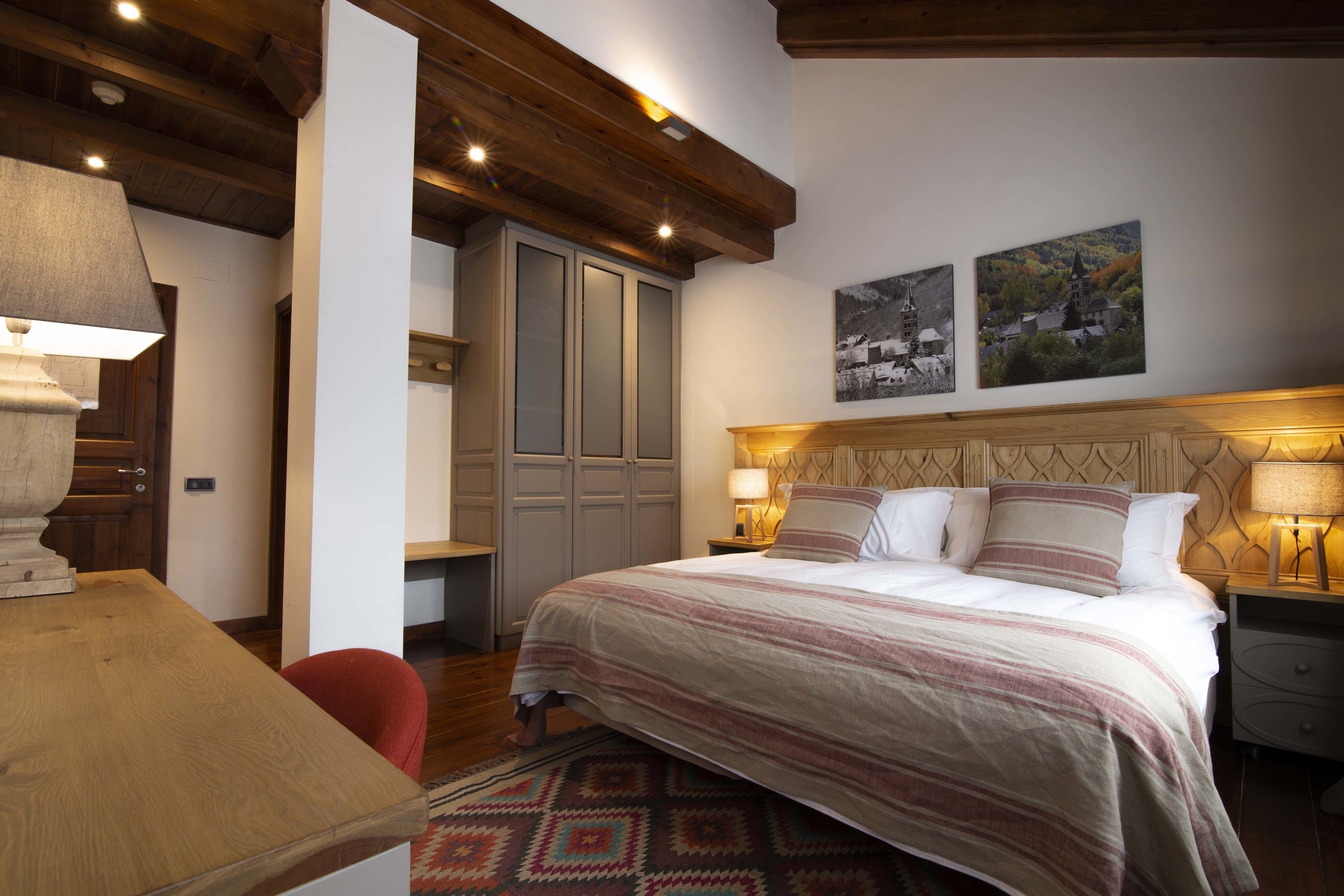A bedroom with a large bed, wooden ceiling beams, a desk, and a chair. There are two wall-mounted lamps and a door with a mirror.