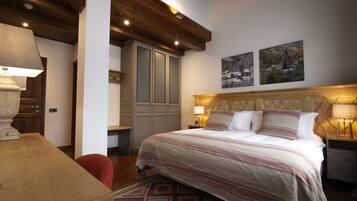 A bedroom with a large bed, wooden ceiling beams, a desk, and a chair. There are two wall-mounted lamps and a door with a mirror.