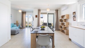 A modern kitchen with a dining table, chairs, and a fruit bowl. There is a sofa and a small table with a lamp in the living area. - 3-bedroom villa close to the beach at Triton SeaView Villas with common poolS (Asprouliani)