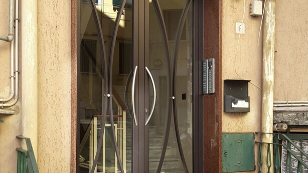 Entrada de un edificio moderno con puerta de vidrio y estructura metálica, una reja metálica verde y unos pocos escalones que conducen a la puerta.
