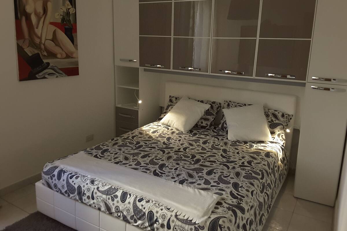Un dormitorio con cama, mesita de noche, lámpara, cómoda y un cuadro en la pared.