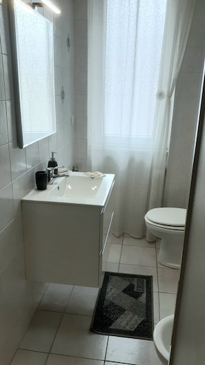 A bathroom with a white sink, mirror, and toilet. There is a window with a curtain, and a black rug on the floor. - CASA DELL'ARTISTA 1 (Salerno)