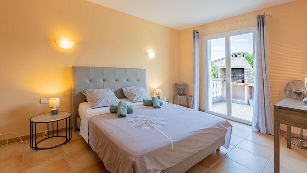 A bedroom with a large bed, two bedside lamps, a small table, and a view of an outdoor grill area through a sliding glass door.