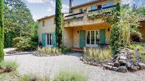 Une maison de style méditerranéen avec une allée en gravier, des volets verts et un mur en pierre.