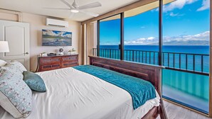 Ein Schlafzimmer mit großem Fenster und Meerblick, Deckenventilator, Bett mit blau-weißer Bettdecke und Nachttisch aus Holz mit Lampe.
