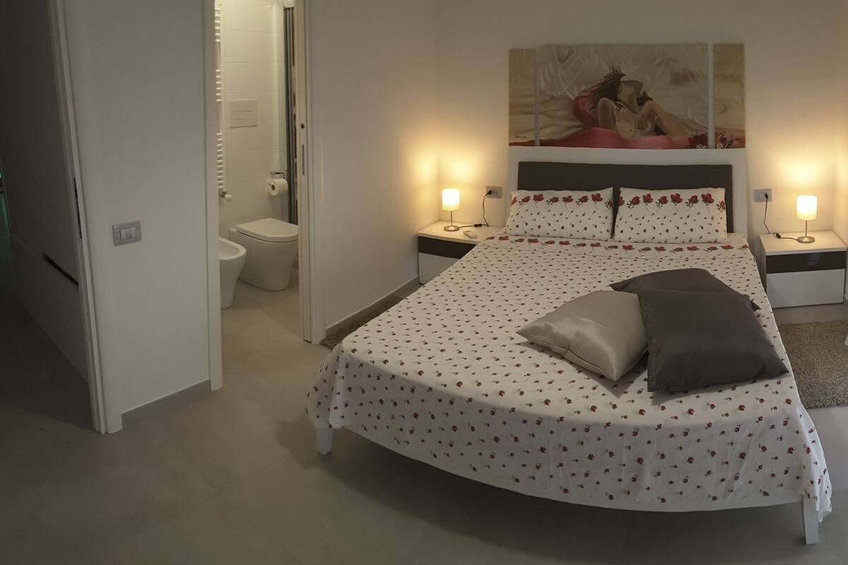 Ein Schlafzimmer mit einem Bett, zwei Nachttischen mit Lampen, einem Bild an der Wand und einem Badezimmer mit Toilette und Waschbecken.
