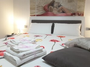 Ein ordentlich bezogenes Bett mit einer blumigen Tagesdecke, ein Stapel Handtücher und ein Kissen daneben.