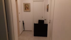 Un couloir étroit, avec une porte au fond, un tableau encadré au mur et un objet noir au centre.
