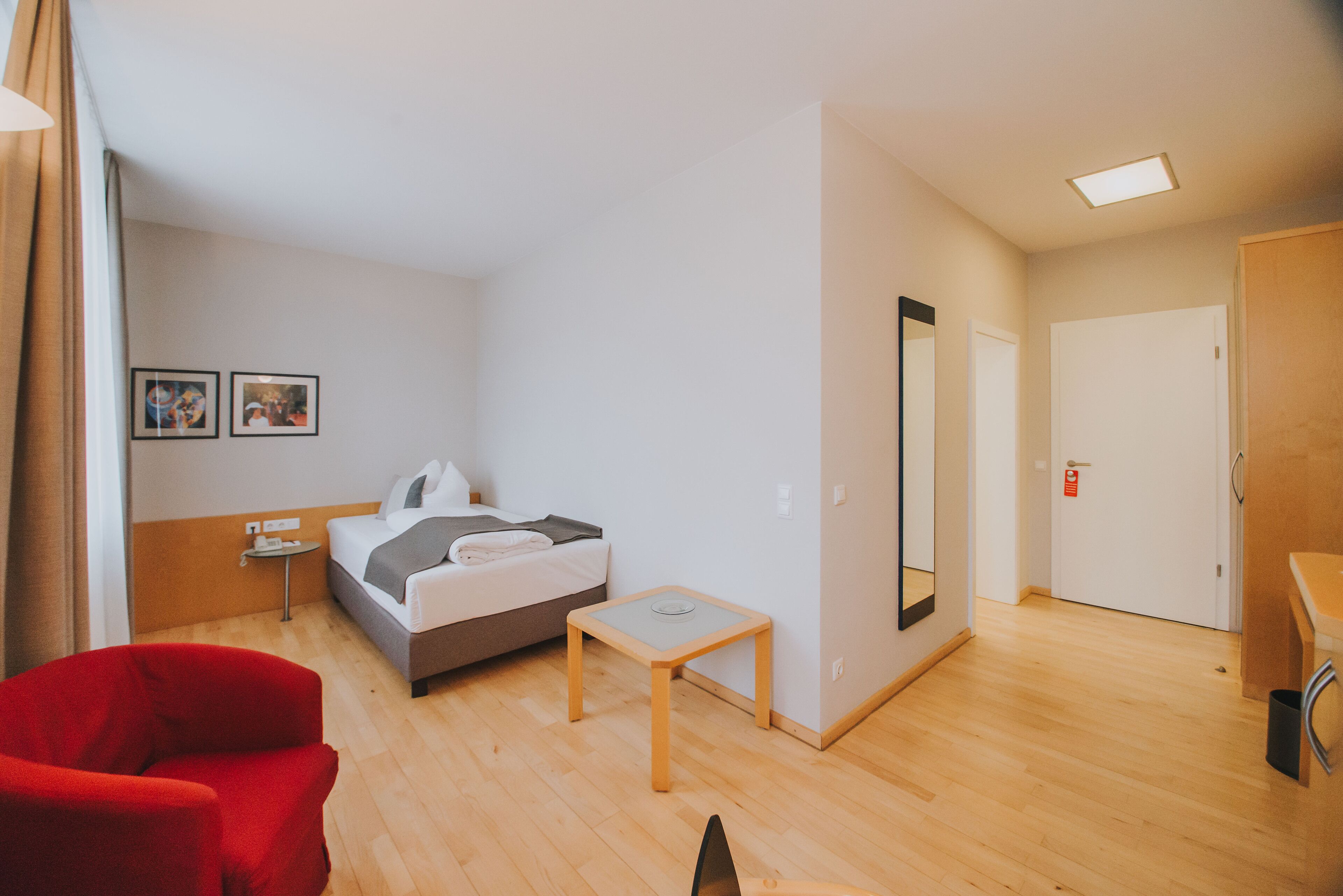 Ein Hotelzimmer mit einem Bett, einem kleinen Tisch, einem roten Sessel, einem Spiegel und einer Tür.