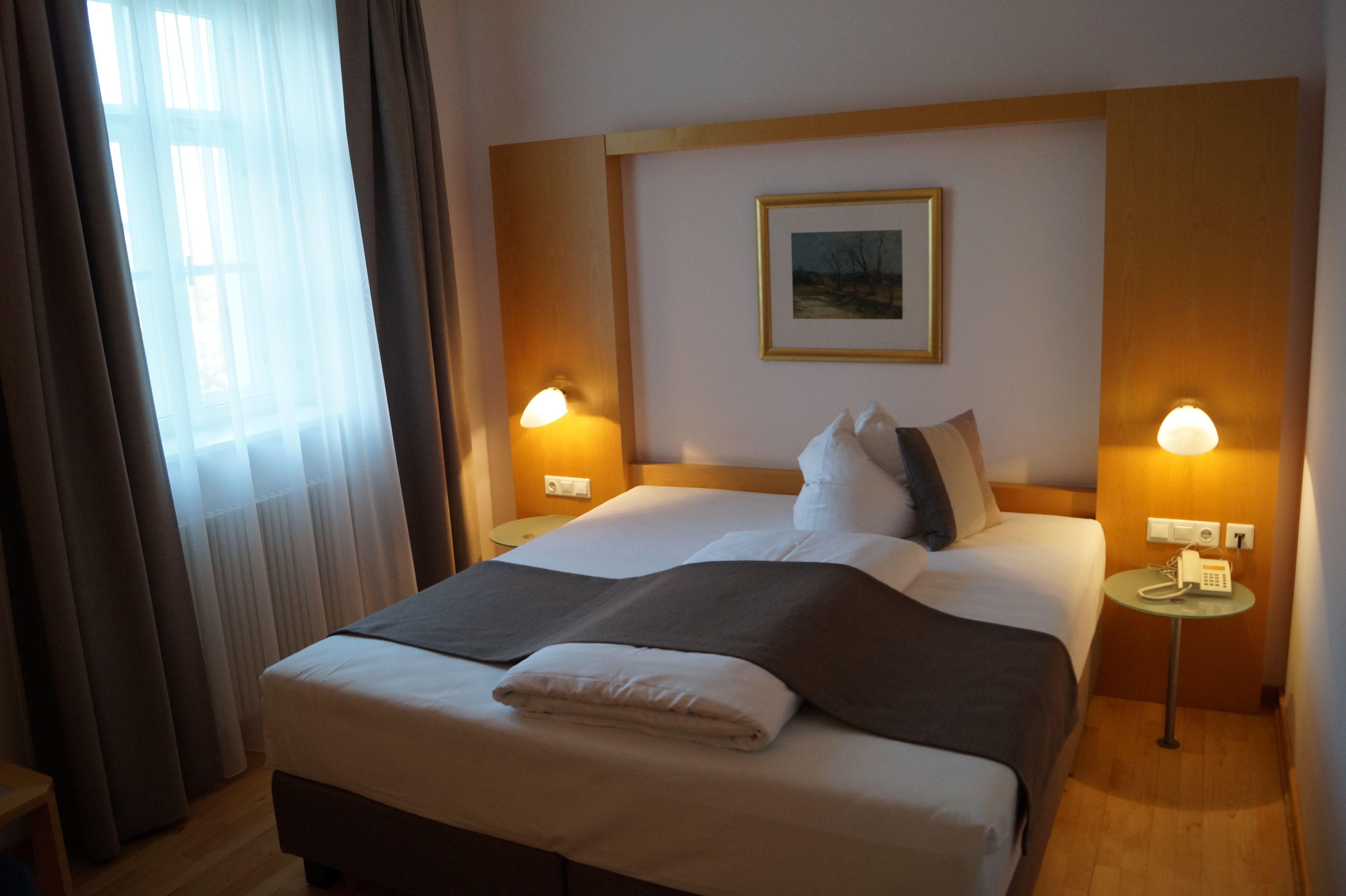 A hotel room with a large bed, two bedside tables with lamps, a painting on the wall, and a window with curtains.