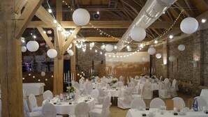 Un salón de banquetes con mesas redondas dispuestas para un evento formal, con manteles blancos y sillas, además de luces colgantes.