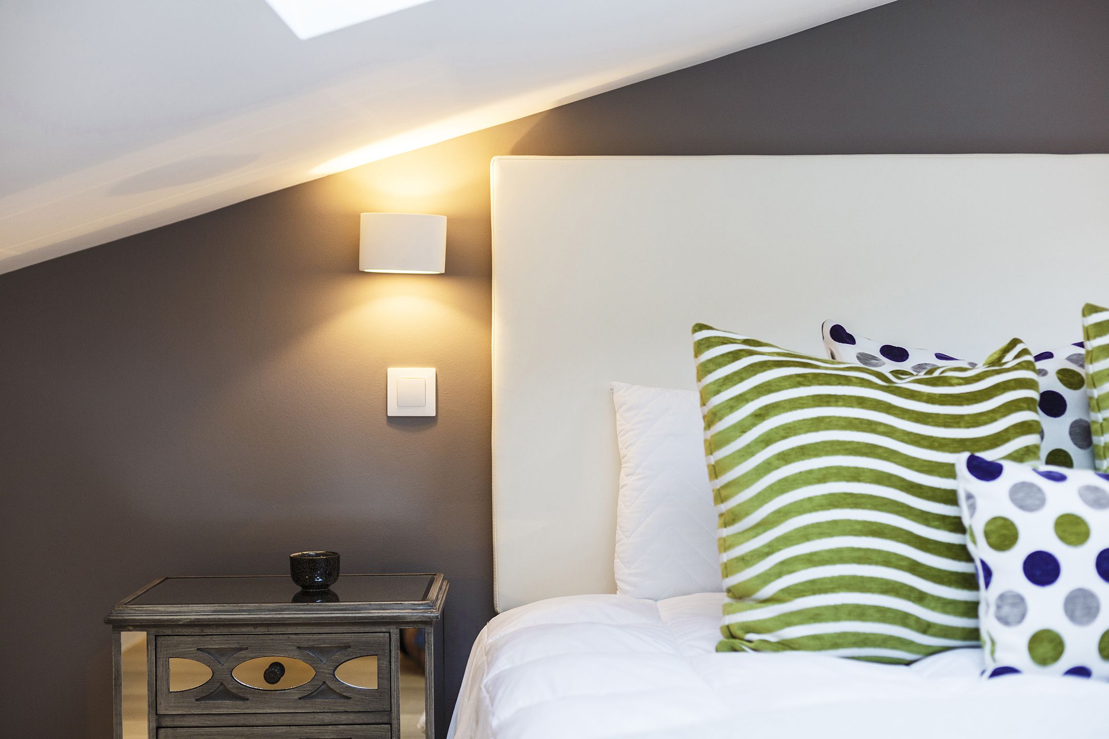 Een slaapkamer met schuin dak, nachtkastje met lamp en een bed met gestreepte kussens.