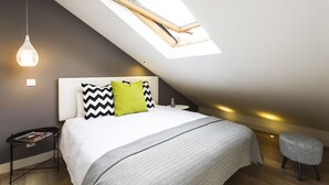 A modern bedroom with a skylight, a bed with patterned bedding, a bedside table with a magazine, and a small stool. - Madalena Downtown Luxury Duplex (Lisbon)