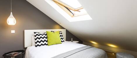 A modern bedroom with a skylight, a bed with patterned bedding, a bedside table with a magazine, and a small stool.