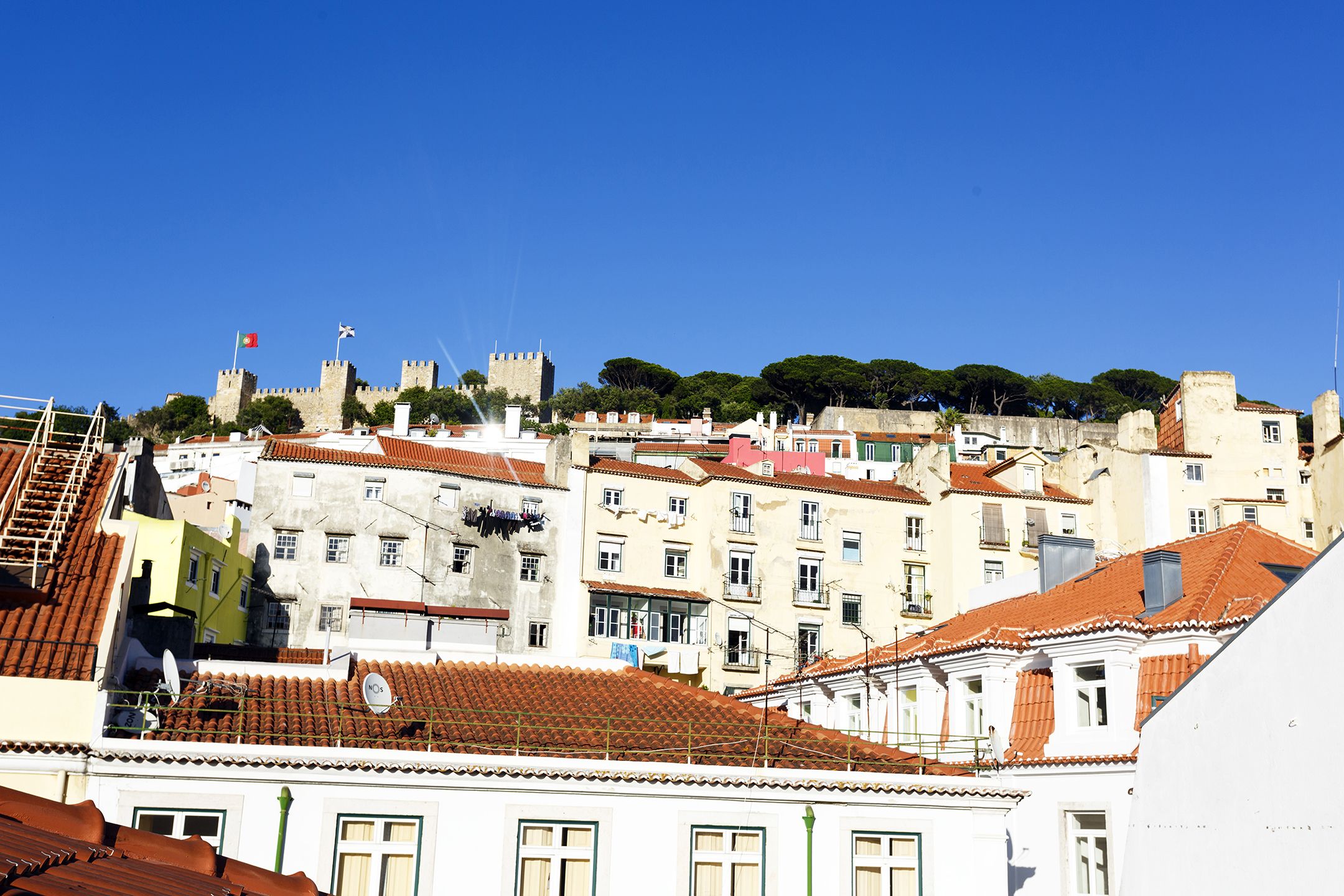 Un paysage urbain d’importance historique, avec une imposante structure ressemblant à un château, des bâtiments aux toits de tuiles en terre cuite et un ciel d’un bleu éclatant.
