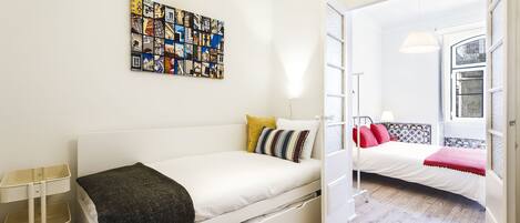 Een moderne, goed verlichte kamer met een wit bed, een klein nachtkastje en een kleurrijk kunstwerk aan de muur.