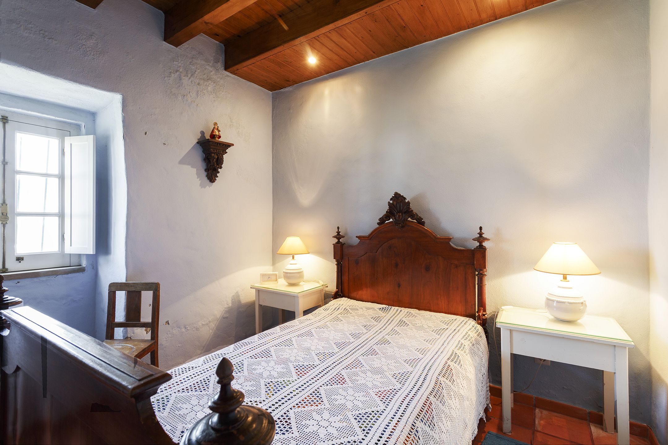 Camera da letto con un letto di legno, due comodini con lampade, una sedia e una finestra con porta aperta.