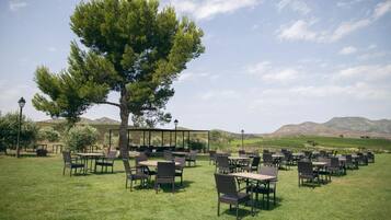 Een terras met tafels en stoelen, omgeven door gras en een grote boom.