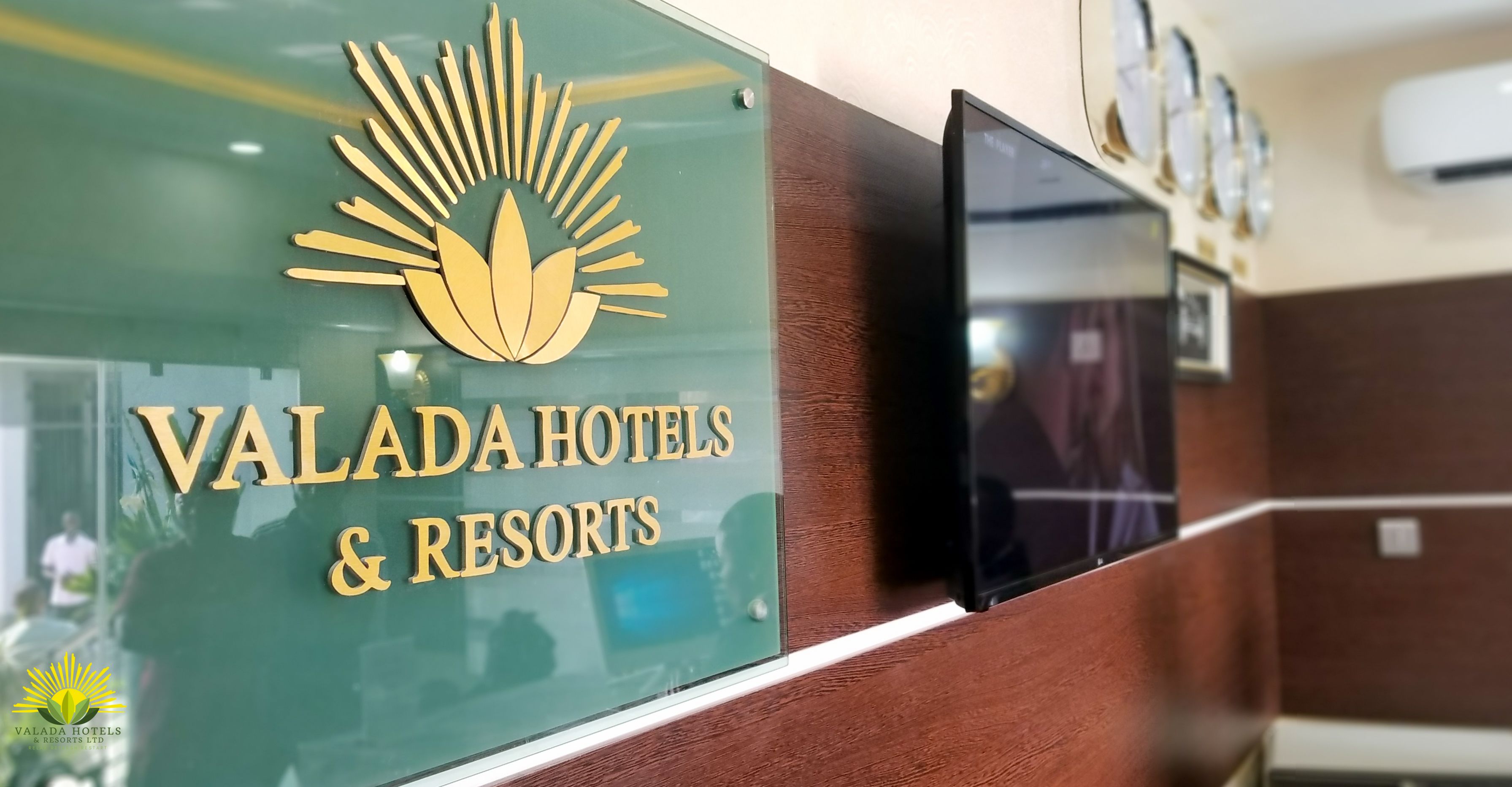 Un comptoir d’accueil avec une enseigne indiquant « Valada Hotels & Resorts », un téléviseur fixé au mur et une horloge.