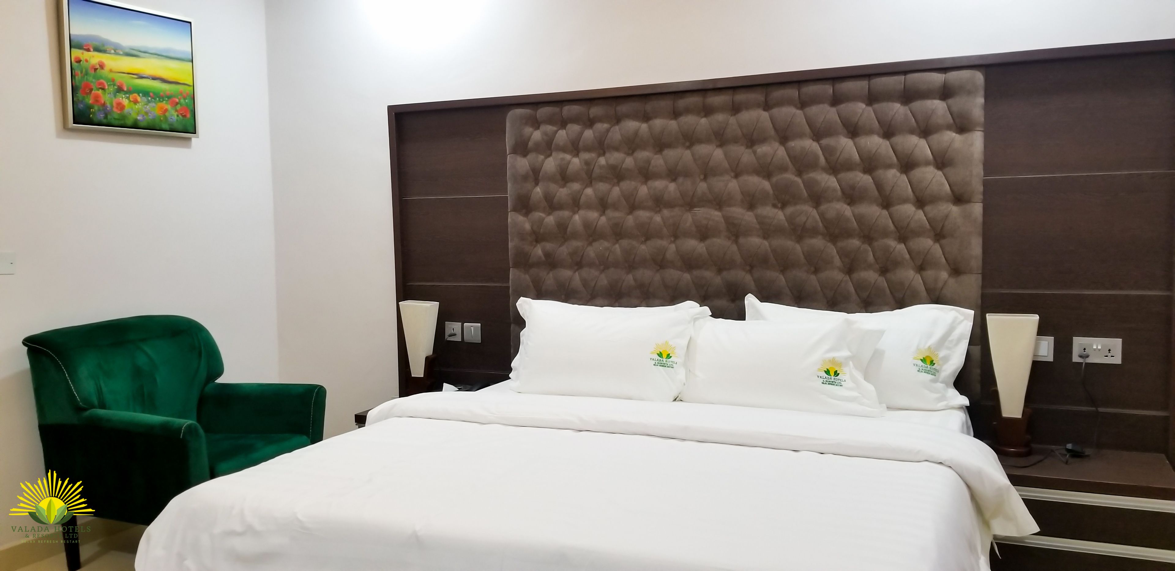 Quarto de hotel com uma cama grande, uma poltrona verde e um quadro na parede.