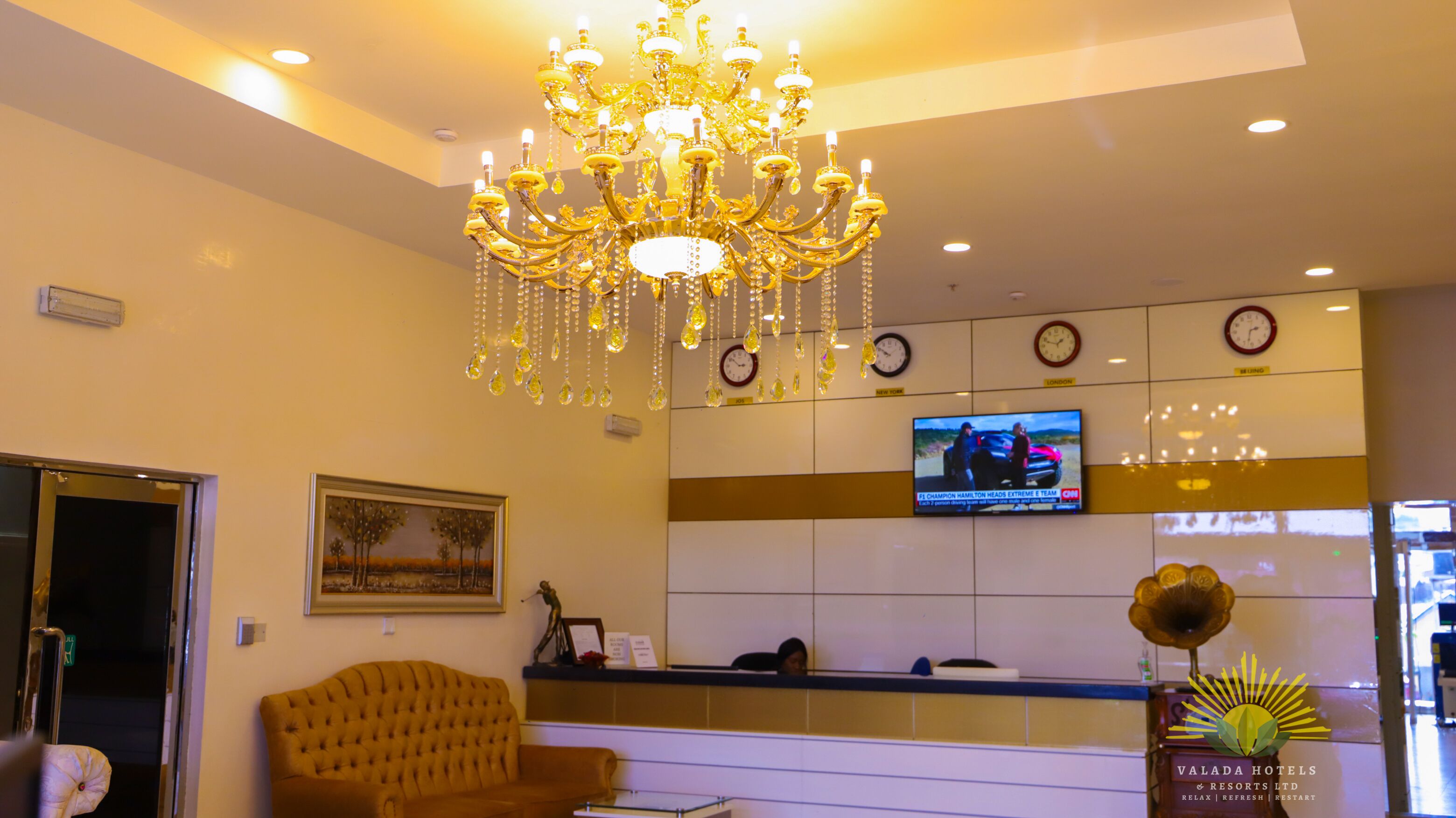 A hotel reception area with a chandelier, a television displaying a news program, a clock, and a framed landscape painting.