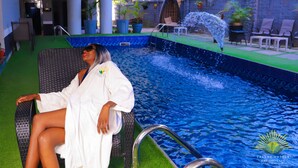 Una persona sentada junto a una piscina con fuente, rodeada de sillas de descanso y plantas en macetas.