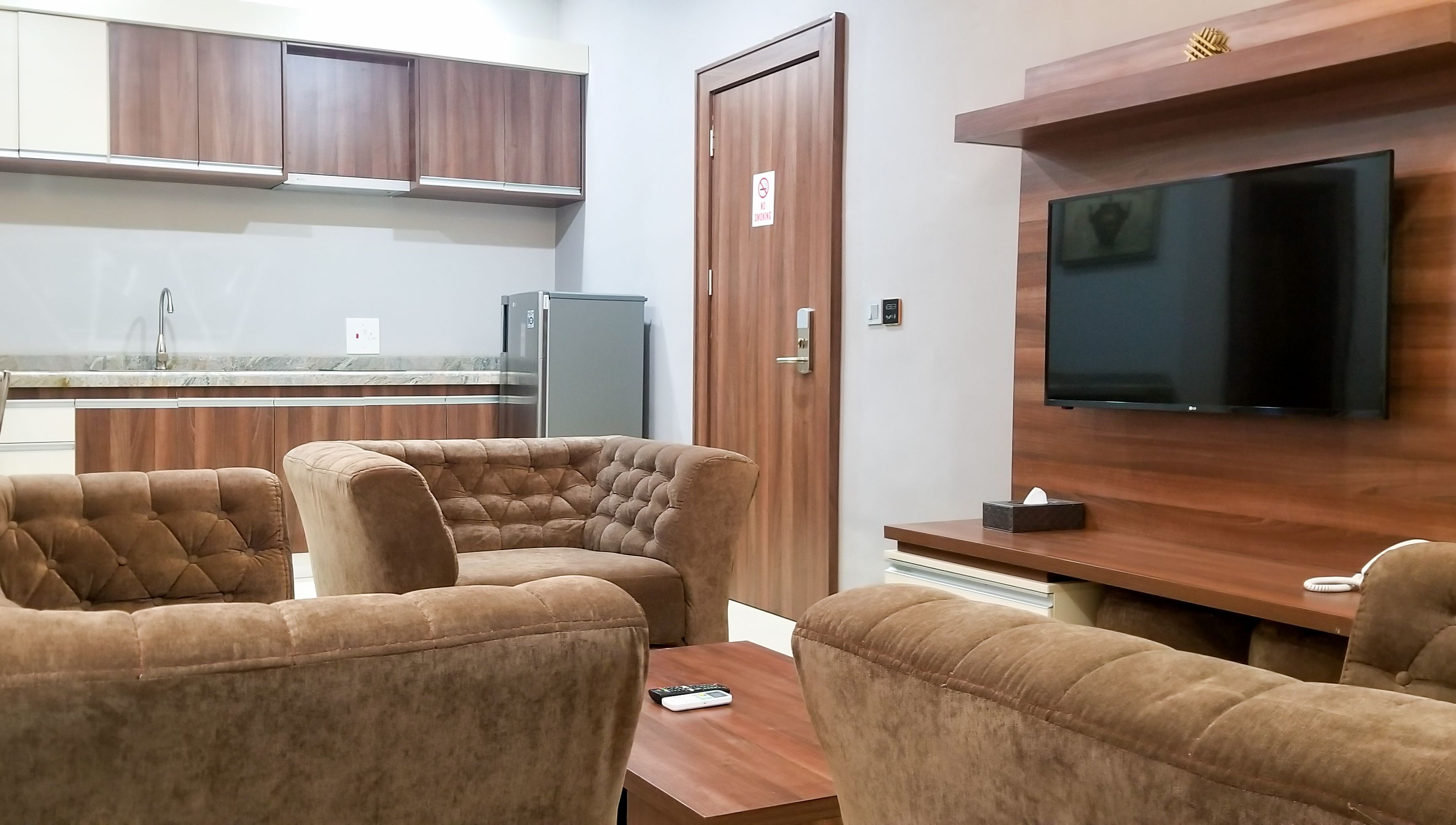 A modern living room with a flat-screen TV, brown upholstered sofas, a wooden coffee table, and a kitchen area with wooden cabinets and a refrigerator.
