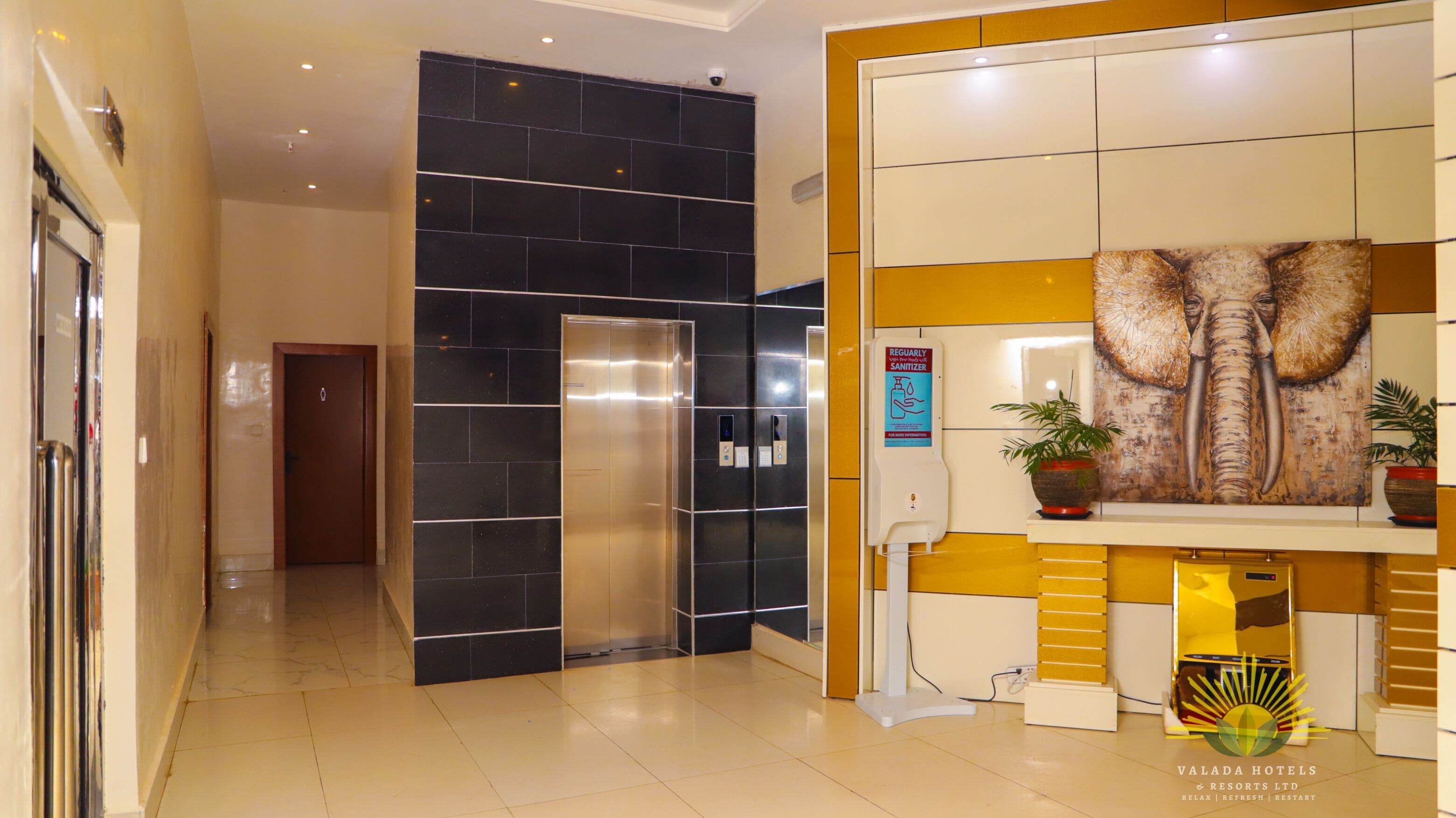 A hotel reception area with a modern elevator, a large elephant artwork, and a sign promoting sanitizer use.