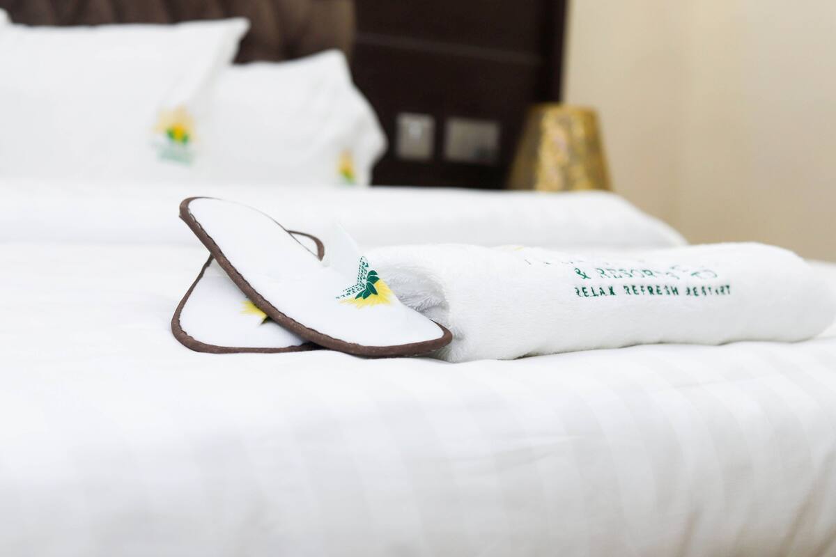 A neatly made bed with white bedding and a pair of slippers with a green logo.