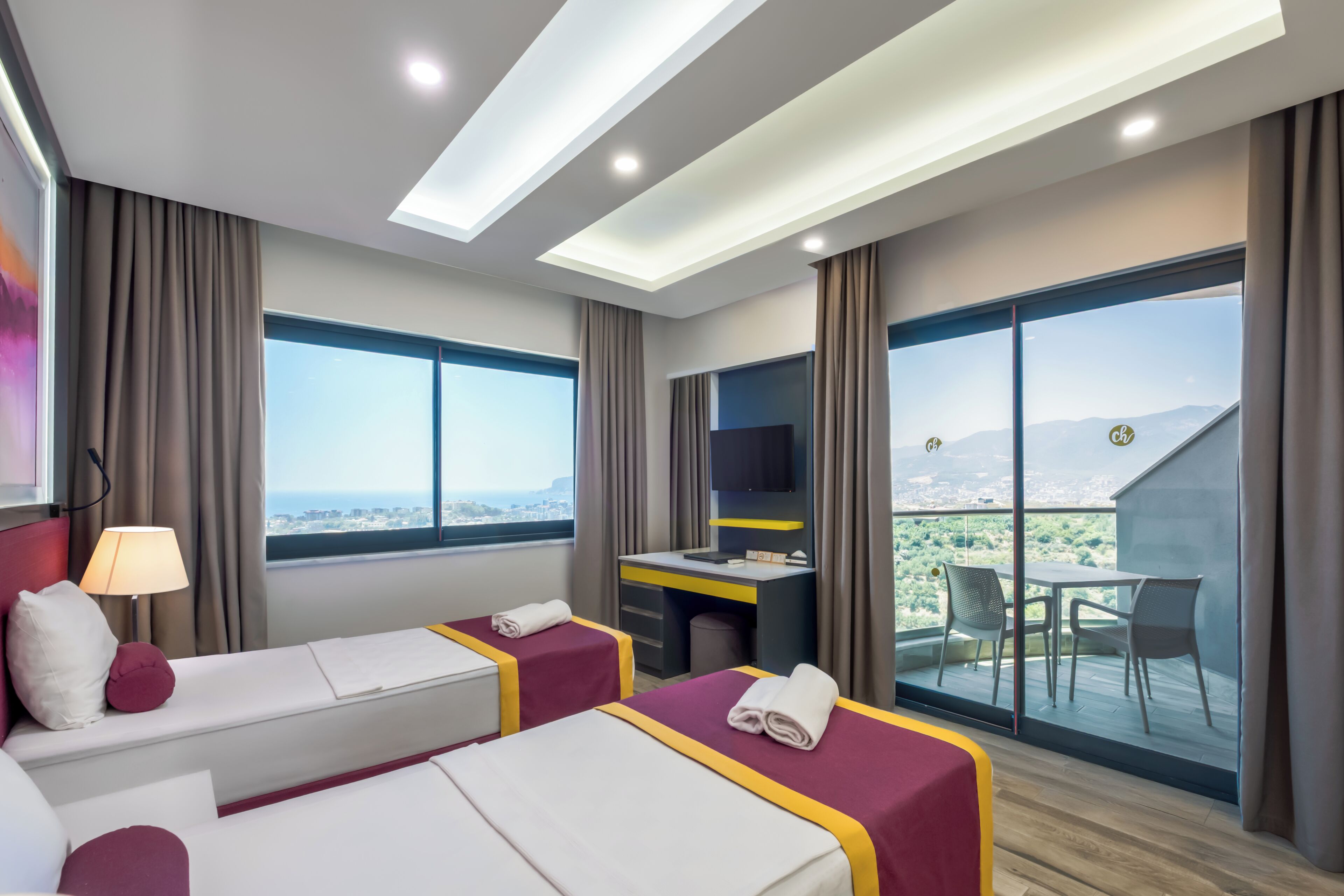 A hotel room with two beds, a desk, a chair, and a balcony with a city view.