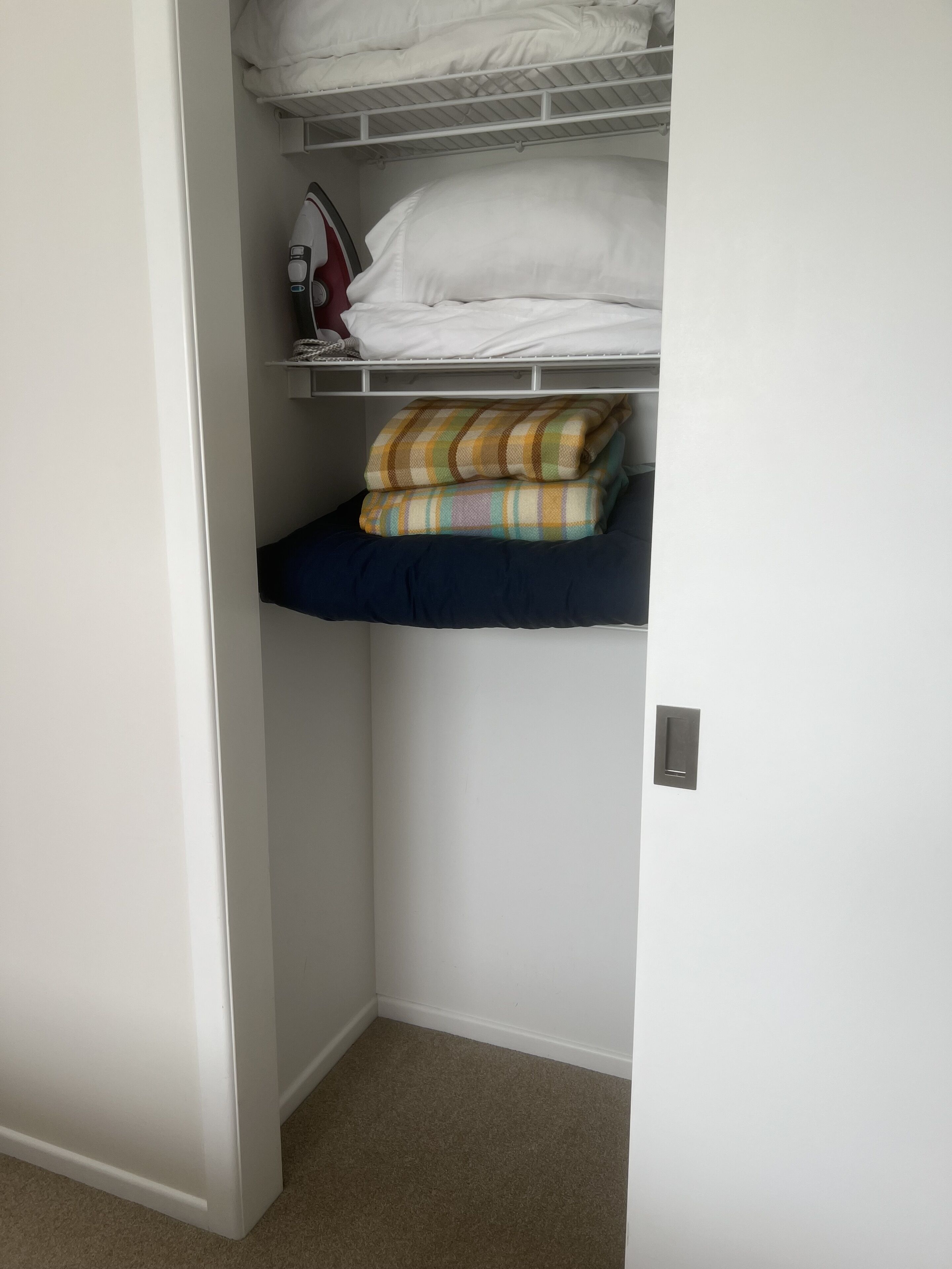 A closet with shelves holding folded clothes, a shelf with a patterned blanket, and a white iron on the top shelf.