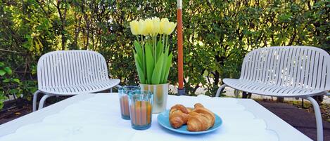 A table with a white tablecloth, two white chairs, a vase of yellow flowers, a metal bucket, two glasses with a beverage, and a plate with croissants.