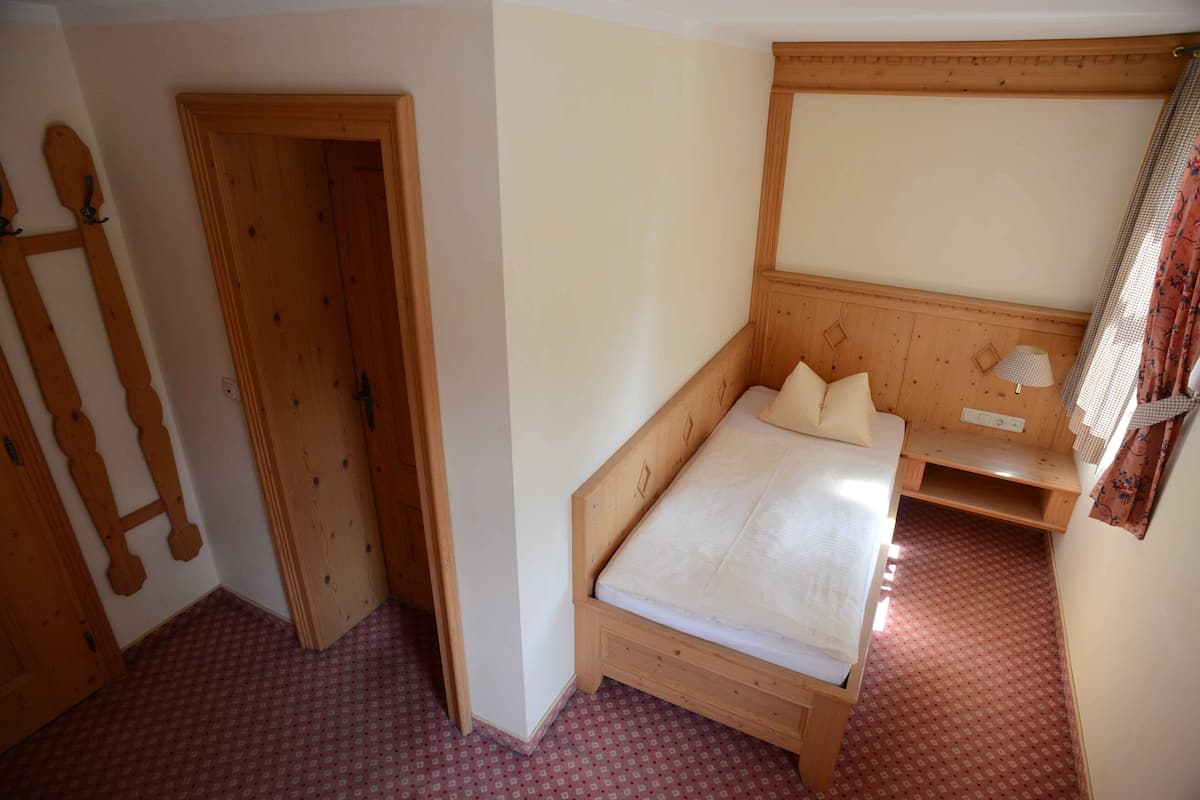 A small bedroom with a single bed, a wooden headboard, a nightstand, and a window with curtains.