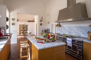 A modern kitchen with a marble countertop, wooden stools, a central island with fresh produce, a stainless steel range hood, and a built-in oven.