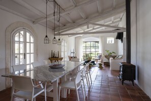 A spacious dining area with a long table and chairs, a rustic stove, and large windows allowing natural light.