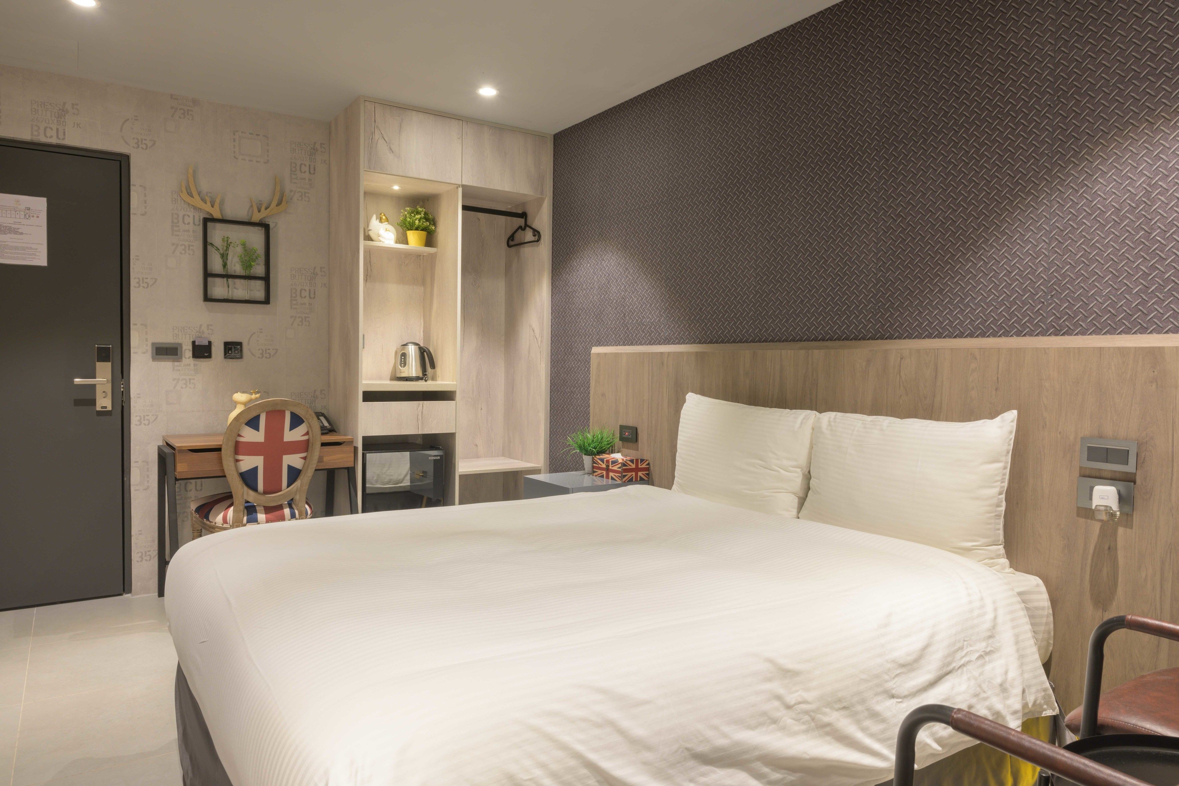 Una habitación de hotel moderna con una cama grande, un armario, un escritorio pequeño con una tetera, un perchero y un estante montado en la pared con plantas.