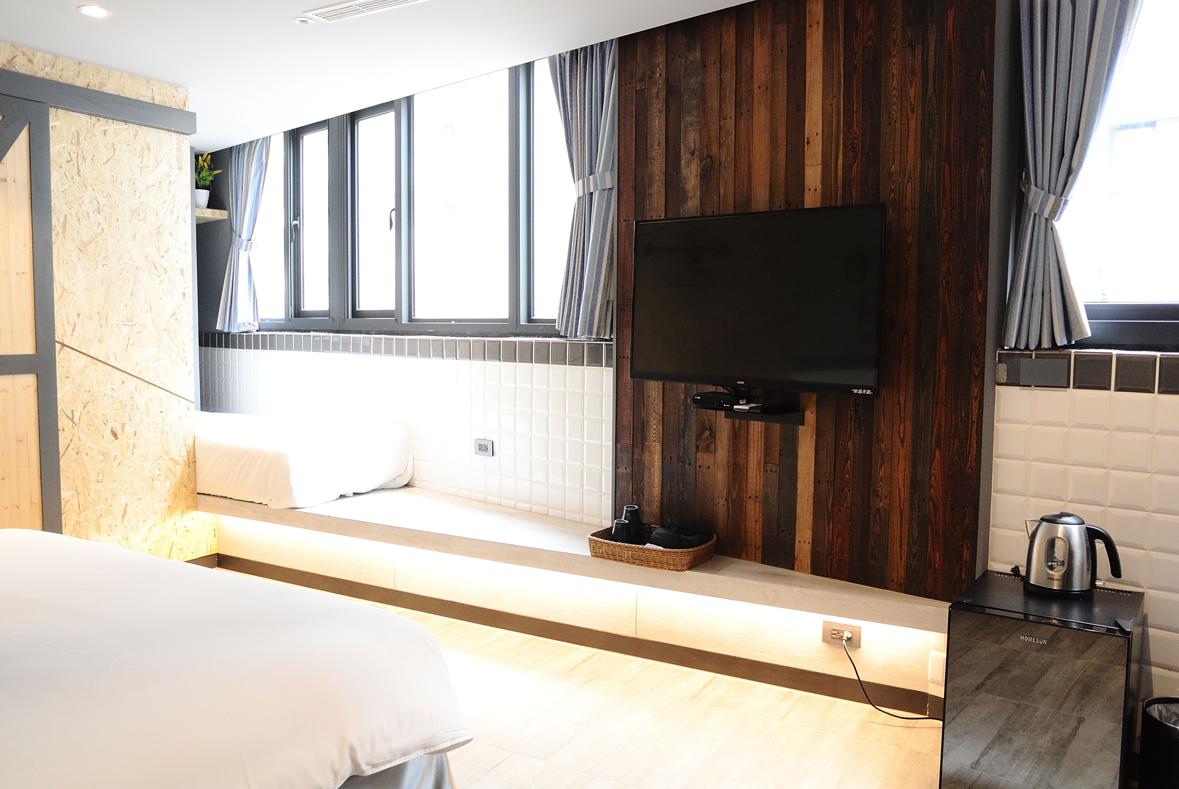 A modern hotel room with a large bed, a TV mounted on a wooden wall, a small kitchen area with a kettle, and a window with curtains.