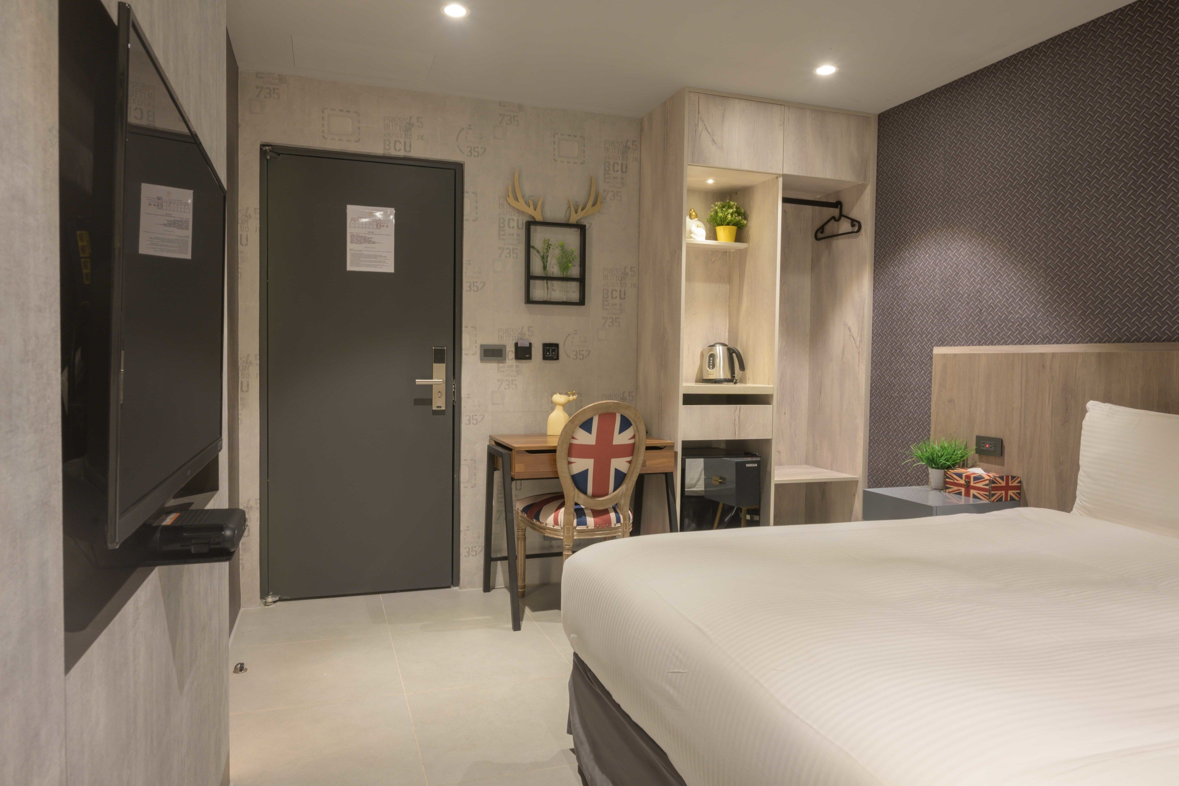 A modern hotel room with a bed, a desk with a chair, a wardrobe, and a door.