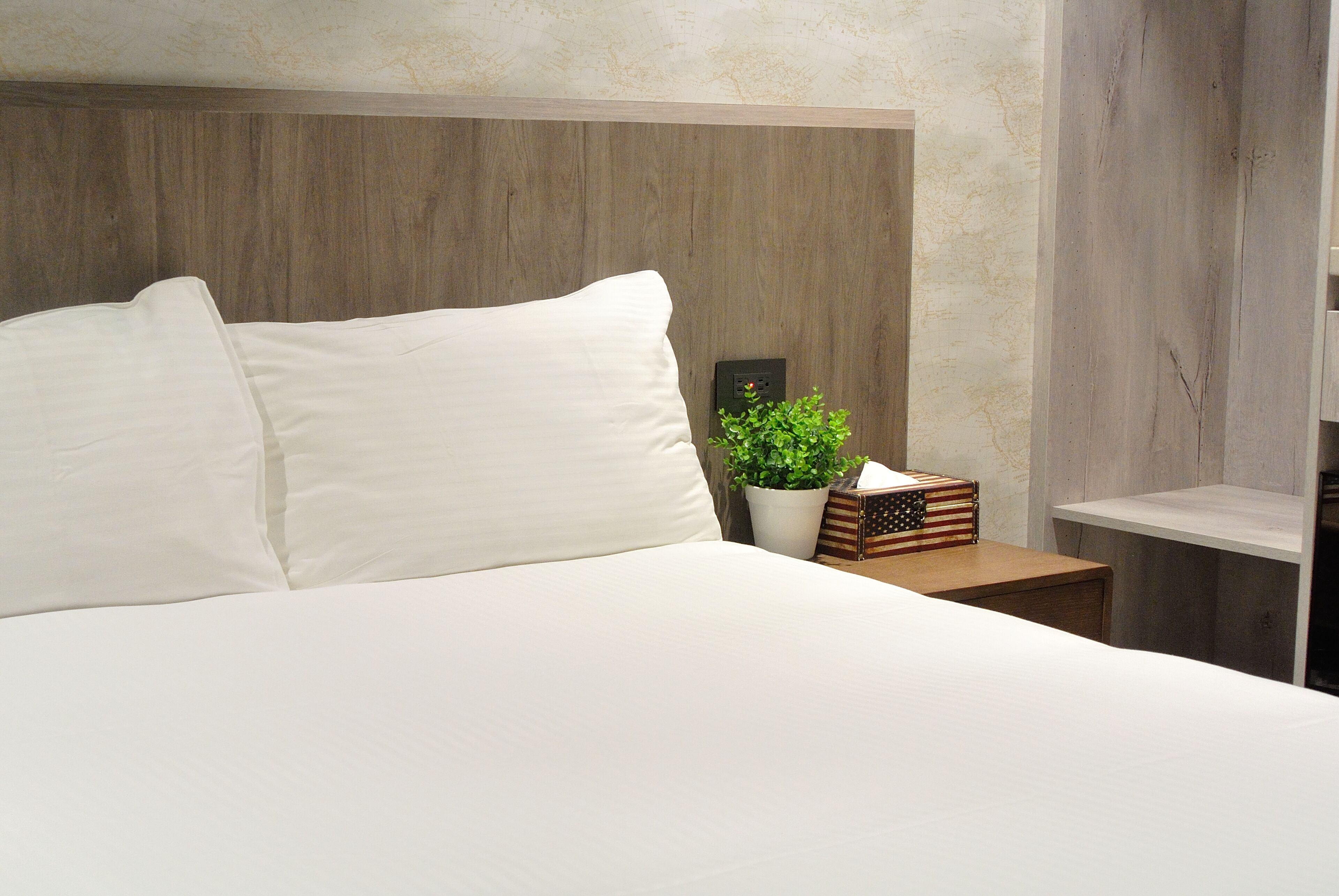 A neatly made bed with white linens, a wooden headboard, a small potted plant, and a nightstand with a lamp and a decorative box.