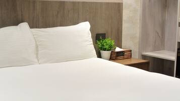 A neatly made bed with white linens, a wooden headboard, a small potted plant, and a nightstand with a lamp and a decorative box.