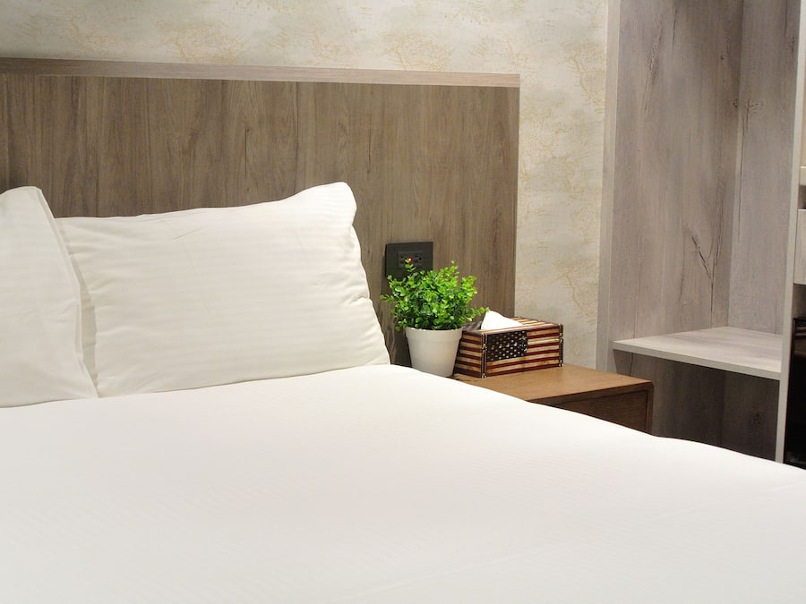 A neatly made bed with white linens, a wooden headboard, a small potted plant, and a nightstand with a lamp and a decorative box.