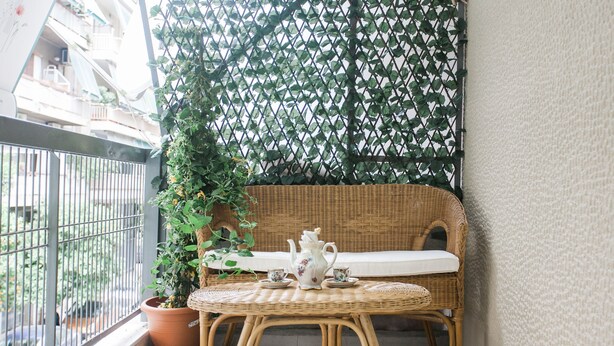 A wicker chair with a cushion, a small table, a teapot, and a cup set on a balcony with a potted plant and a decorative fence. - Spacious Safe Apartment Walk Acropolis (Athens)
