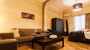 A modern hotel room with a wooden floor, a sofa, a dining table with a plate of fruit, a bed with a patterned bedspread, and a painting on the wall. - Spacious Safe Apartment Walk Acropolis (Athens)