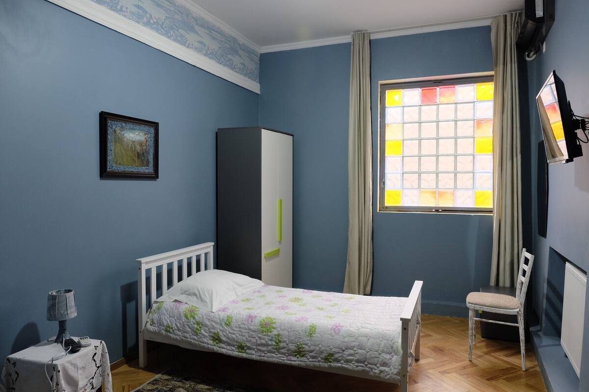 A bedroom with a bed, a chair, a wardrobe, a window with curtains, and a painting on the wall.
