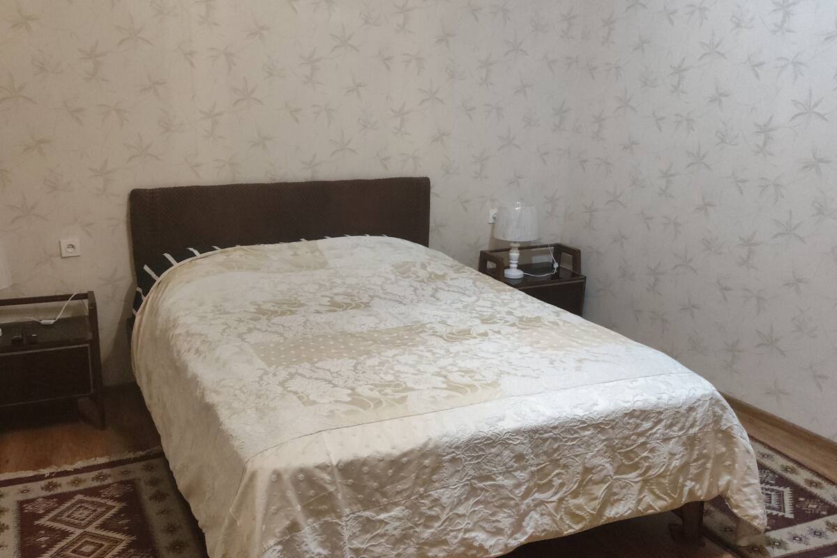A small, neatly made bedroom with a single bed, a nightstand, a lamp, and a bedside table with a lamp. The room has patterned wallpaper and a patterned rug on the floor.